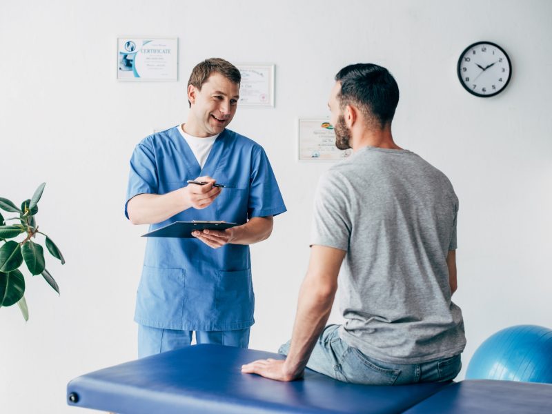 smiling-physiotherapist-with-diagnosis-and-pen-gesturing-near-patient-in-hospital.jpg